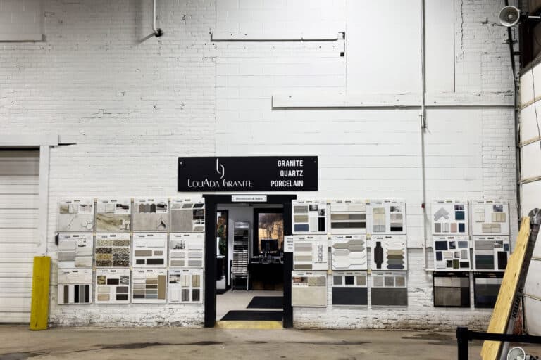 A showroom entrance with a sign reading “Louada Granite” and wall displays of granite, quartz, and porcelain samples. The space has white brick walls and industrial decor.